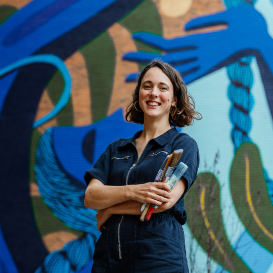 Zoe (artist) smiling at the camera with her arms crossed, holding paintbrushes in one hand, a mural with hues of blue behind her.